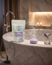 A stand-up pouch of Lavender Organic Bath Bombs sits on a marble bathroom counter beside a glass of lavender water and a floating bath bomb with rising steam.