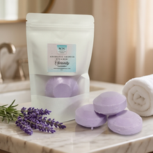 A white stand-up pouch of RDC Organic Aromatic Shower Stones, labeled Lavender, sits on a marble bathroom counter next to three round lavender-colored bath stones, a sprig of lavender, and a rolled white towel.