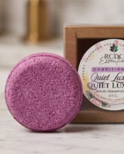 A round purple solid shampoo bar in front of its kraft paper box packaging on a light marble surface. The label on the box reads 'RCDC Essentials Quiet Lux Quiet Lux'.