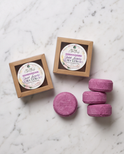 Three round purple bath salts are arranged on a white marble surface next to two small kraft paper boxes with round white labels. The labels read 'Enchanting Quiet Luxury' and the brand appears to be 'The Art of Bathing'.