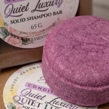 A round purple solid shampoo bar in the foreground with two round labels behind it reading 'Quiet Luxury Conditioning Solid Shampoo Bar 65 G'.