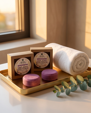 Two small brown gift boxes with round labels sit on a gold tray beside two purple bath soaps and a rolled white towel, styled near a window with warm sunlight and a eucalyptus sprig.