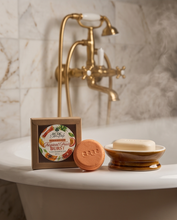 A boxed and wrapped bar of soap sits on a white bathroom counter in front of a brass faucet and marble wall. The round soap in front is terracotta-colored with the brand name 'gojo' embossed on it, and the square box shows a colorful label reading 'Peach & Mango Burst'.