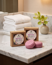 Two pink bath soaps in round pink bars with kraft paper boxes, displayed on a white marble countertop with folded white towels and a small vase of green leaves in the background.