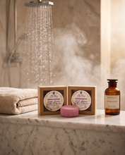 Two boxed bath products and a pink bar of soap arranged on a marble counter in a bathroom setting with a shower head spraying water and steam in the background. A folded beige towel and an amber bottle are also visible.