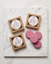 Four round pink bath salts are arranged next to two small kraft paper boxes containing round labels, all placed on a white marble board. The labels read 'St.