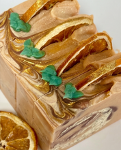 A close-up of a rectangular bar of artisanal soap with a light tan base, metallic gold swirls, dried orange slice inclusions, and small green leaf accents, photographed on a clean white background.