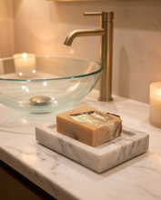 A rectangular bar of marbled soap in warm tan and cream tones sits on a white marble soap dish on a bathroom counter, with a glass bowl, brass faucet, and lit candles in the background.