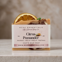 A bar of goat's milk soap from RCDC Essentials, Citrus Pomander scent, displayed in its cardboard packaging with a dried orange slice on top, set on a light stone surface against a neutral marble background.