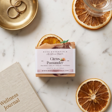 A bar of RCDC Essentials Citrus Pomander goat milk soap is wrapped in kraft paper and displayed on a white marble surface with dried citrus slices, a gold tray with rings, and a journal.