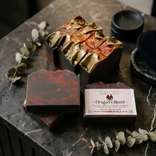 A styled product shot of artisanal soap bars and a branded box on a dark marble surface. The soaps have deep reddish-brown and metallic gold marbled patterns, with one bar sliced open to show the layered texture.