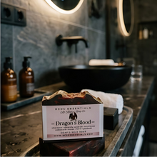 A bar of soap from BCDC Essentials 'The Cat & Mouse Co.' labeled 'Dragon's Blood' sits on a bathroom counter in front of a blurred modern vanity with a black vessel sink and illuminated mirrors.