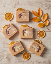 Five rectangular bars of soap with a tan base and marbled swirls of cream, brown, and dark plum, arranged on a light wood surface with dried orange slices and orange leaves as props.