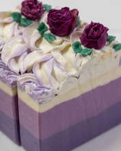 A close-up of a rectangular layered soap bar decorated with piped lavender and white frosting swirls, topped with three dark purple rose decorations and green leaves, on a plain light background.