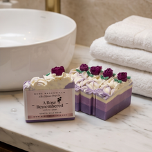 Two decorative goat's milk soaps on a marble bathroom counter next to a white sink and folded white towels. The soap bar in front has a paper label reading 'A Rose Remembered' by MCDC Essentials, and both soaps feature purple and cream layers topped with white piped frosting and purple rose decorations.