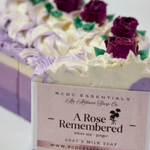 A close-up of a rectangular bar of handmade soap with piped white and lavender swirls and purple rose decorations on top. The front face has a paper label reading 'RCDC ESSENTIALS The Artisan Soap Co.
