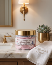 A jar of RCO Essentials Whipped Soap Scrub labeled 'Mermaid Dreams' sits on a marble bathroom counter beside a folded white towel embroidered 'Mermaid's Touch,' with a gold sconce, plant, and brass faucet in the background.