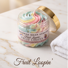 A clear jar of whipped soap scrub labeled Fruit Loopin' by KCdCeEssentials, filled with pastel whipped soap swirls, set on a white marble surface with a gold lid leaning behind it and a folded white towel nearby.