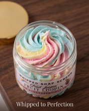 A clear jar of whipped soap scrub with pastel blue, pink, yellow, and white swirled scrub visible through the lid, labeled Fruit Loopin' by RCD Essentials. The jar sits on a dark wood surface with a gold-toned lid resting beside it.