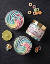 Three jars of whipped soap scrub arranged on a dark slate background, including two open round jars showing pastel swirls and one closed jar with a gold lid. A lime slice and cereal ring props are placed around the products.
