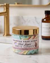 A clear jar of Fruit Loopin Whipped Soap Scrub with a shiny gold lid and a pink label, set on a white marble countertop with a gold faucet and an amber bottle in the background.