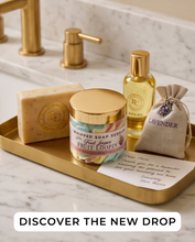 A styled bathroom scene with a gold tray holding a bar of soap, a jar of whipped soap scrubs, a bottle of oil, a lavender drawstring pouch, and a card on a marble surface. A large banner at the bottom reads 'DISCOVER THE NEW DROP'.