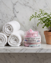 A jar of whipped soap scrub labeled 'Candy Pearls' sits on a white marble shelf beside three rolled white towels and a potted green plant. The jar contains pink whipped soap with blue and white swirls.