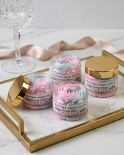 Four jars of whipped soap scrub labeled 'The Candy Cloud' arranged on a gold tray with a marble surface, alongside a champagne-colored ribbon and a crystal glass in the background.