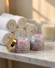 Two jars of Whipped Soap Scrub in Candy Cloud Pink and Blue scents on a marble bathroom counter, with rolled towels and a lit candle in the background.