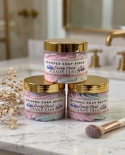 Three jars of Whipped Soap Scrub in the 'Candy Cloud' scent, stacked in a pyramid on a white marble countertop with a makeup brush and dried flowers nearby. The background shows a softly blurred bathroom setting with gold fixtures.