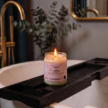 A lit candle in a clear glass jar sits on a black wooden bathtub caddy, placed over a white tub in a styled bathroom setting with a gold faucet, mirror, and eucalyptus decor.