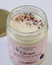 A close-up of a glass jar candle with cream-colored wax topped with multicolored confetti, labeled 'Signature Zodiac Collection Scorpio' by RCDC Essentials, with its gold lid resting in the background.