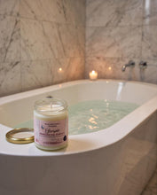 A jar candle labeled 'Cherpie Unearth the Sulfur' sits on the edge of a white bathtub with marble tile walls and a lit candle in the background.