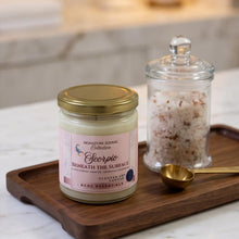 A jar candle labeled 'Signature Zenith Collection Scorpio Beneath the Surface' sits on a wooden tray next to a glass jar of bath salts and a small gold spoon, set on a marble surface with a blurred bathroom background.