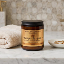 A soy wax candle in an amber glass jar with a black lid and a tan label reading 'Ginger & Spice' by RCDC Essentials Home Collection, styled on a marble countertop with a folded towel and gold jewelry in a bathroom setting.