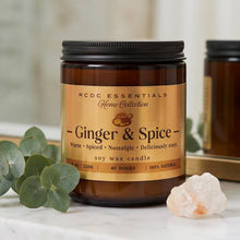 An amber glass soy wax candle jar labeled RCDC Essentials Ginger & Spice, styled on a white marble surface with eucalyptus leaves and a crystal, with a second candle blurred in the background.