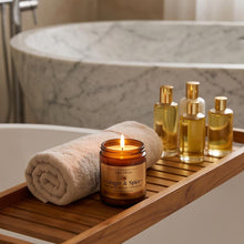 A lit amber glass jar candle labeled Ginger & Spice sits on a wooden bath tray draped over a bathtub, alongside a rolled beige towel and four gold-toned bottles in a bathroom setting.