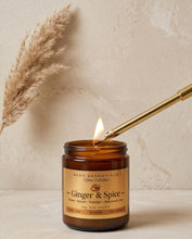 An amber glass jar candle labeled 'Ginger & Spice' by HCDC Essentials Home Collection, lit with a gold metal lighter against a warm beige textured background with dried grass.