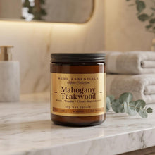 An amber glass soy wax candle jar with a black lid and a tan label reading 'RCCC Essentials Home Collection Mahogany Teakwood' sits on a white marble countertop in a softly blurred bathroom setting with folded towels and greenery.