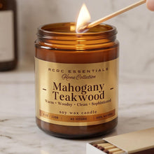 An amber glass soy wax candle jar labeled 'RCDC ESSENTIALS Home Collection Mahogany Teakwood' with a lit match being held near the flame, on a marble surface with a matchbox nearby.