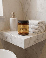 A jar candle labeled 'Mahogany Teakwood' by Wood Essentials sits on a marble floating shelf next to a white soap dispenser and a stack of folded white towels.