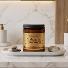 A jar candle labeled 'BCDC Essentials Home Collection - Mahogany Teakwood' sits on a white tray with stones, set on a marble countertop in a warm, modern bathroom setting.