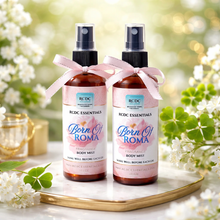 Two RCDC Essentials Born of Roma fragrance body mist bottles on a decorative tray with flowers in the background.