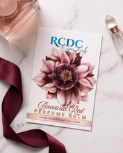 A flat-lay of an RCDG Essentials perfume balm card featuring a watercolor flower design, styled on a white marble surface with a burgundy ribbon and perfume bottles.