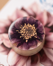 A close-up of a round, clear resin coaster or cup featuring a detailed dark purple and black flower design with a metallic gold stamen center, resting on a blurred pink floral background.