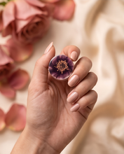 A hand with a pale pink manicure holds a small circular clear dome containing a preserved purple flower bud with a yellow-orange center, against a soft beige background with blurred pink rose petals.