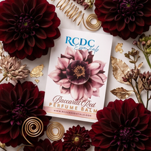RCDC Essentials 'Buccali Red' Perfume Balm card, styled in a flat lay with deep burgundy dahlias, gold foil accents, and dried botanicals on a white marble background.