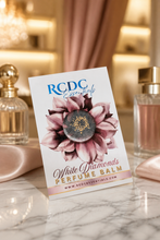 RCDC Essentials  Diamonds Perfume Balm packaging card standing on a marble surface, with perfume bottles and soft pink fabric in a warm, softly blurred boutique setting.