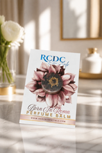 RCDC 'Born In Roma' Perfume Balm in a white stand-up card packaging, featuring a watercolor pink flower and a dark brown balm piece in the center, set on a marble surface with soft sunlight and blurred decor in the background.