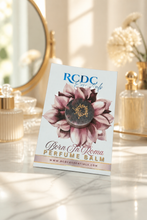 RCDC Essentials 'Born in Roma Perfume Balm' card standing upright on a marble surface, with perfume bottles and a mirror blurred in the background.